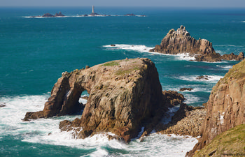 Longships Enys Dodnan This landscape photograph depicts Longships Enys Dodnan, a prominent natural rocky arch along the coast of Cornwall in the United Kingdom. Taken in the late morning during the early spring season, the image captures the rugged cliffs and dynamic coastal scenery with the Atlantic Ocean waves breaking against the rocks. In the background, the Longships Lighthouse is clearly visible, standing on a small island and serving as a landmark for this stretch of the Cornwall coast. The scene highlights the unspoiled nature and dramatic cliffs characteristic of the region.